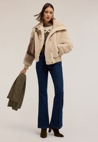 Veste en fausse fourrure beige avec un large col, des poignets côtelés, de gros boutons ; portée par-dessus un pull à motif et un jean bleu évasé, tenant une écharpe verte.