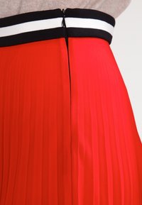 Jupe plissée rouge avec une taille noire à rayures blanches et une fermeture éclair dissimulée au dos. Texture lisse et couleur vive.