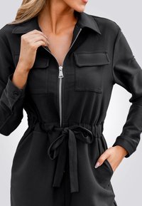 Combinaison noire à manches longues, fermeture éclair à l'avant, deux poches poitrine, taille élastique avec lien, tissu lisse et coupe décontractée.