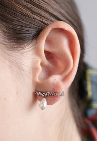 Sudraba auskari ar "Westwood" uzrakstu un asaras formas pērli, kas karājas no vienas puses, valkāti uz auss. 