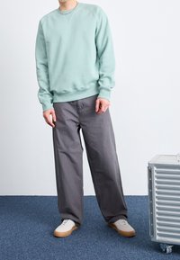 Man med en ljusgrön sweatshirt, lösa gråbyxor och vita sneakers som står på en blå matta bredvid en silverribbad resväska.