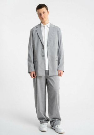 Młoda osoba stojąca w luźnym szarym garniturze, białej koszuli i białych trampkach na jednolitym jasnym tle.