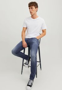 Jean en denim bleu foncé avec une coupe slim, associé à un t-shirt blanc à col rond uni et des baskets noires, assis sur un tabouret en métal.