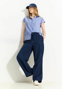 Camicia azzurra chiaro a maniche corte con bottoni abbinata a pantaloni larghi blu navy e sneakers bianche. L'outfit è completato da un cappellino blu navy.