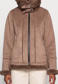 Brun shearlingjacka med dragkedja framtill, pälsfodrad krage och manschetter. Har vertikal sömnad och sidofickor, mjuk textur.