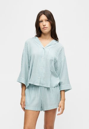Jeune femme portant une chemise boutonnée à rayures verticales bleu clair et blanc et un short assorti sur un fond uni clair.