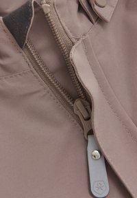 Ljusbrunt tyg med en dragkedja och grå dragflik samt tryckknappsstängning. Slät textur med sömdetaljer nära dragkedjan.