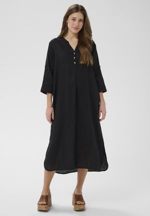 Fiatal nő, aki laza fekete midi ruhát visel háromnegyedes ujjakkal és barna platform szandált, egyszínű háttér előtt áll.