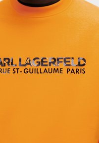 Oranssi puuvillainen t-paita, jossa on musta graafinen printti "CARL LAGERFELD RUE ST-GUILLAUME PARIS." Pyöreä kaula-aukko, lyhyet hihat, sileä pinta.