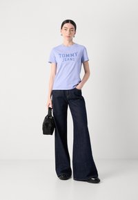 Šviesiai violetinė marškinėlio su užrašu "TOMMY JEANS" tamsiai mėlyna spalva, derinami su tamsiais plačiomis džinsinėmis kelnėmis ir mažą juodą rankinę.