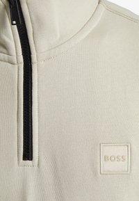 Pullover in pile color beige chiaro con mezza zip nera, caratterizzato da un patch quadrato del logo in beige chiaro. Tessuto liscio, dettagli delle cuciture minimi.