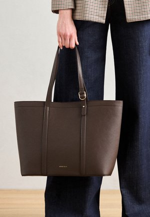Sac fourre-tout en cuir marron avec deux poignées, présentant une texture lisse, des coutures verticales, et un accent en logo doré en bas.