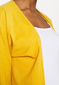 Gul, strukturerad cardigan med lös passform och öppen front, bärs över en vit topp. Vertikala ränder synliga på tyget.