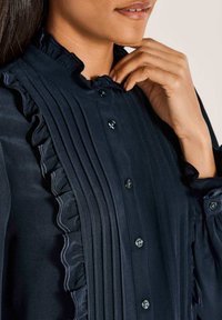 Navybluse mit Rüschung am Kragen, vertikalen Falten und Knopfverschluss mit runden, dunkelblauen Knöpfen. Glatter, strukturierter Stoff.