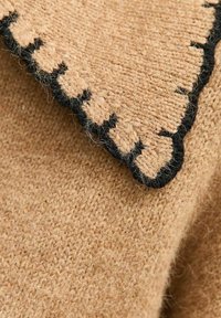 Tessuto a maglia beige con una superficie texturizzata, caratterizzato da un bordo scallop ricamato in nero sul colletto per un contrasto.