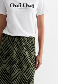 T-shirt blanc avec le texte noir "Oui Oui Mademoiselle", associé à une jupe verte avec un motif en zigzag noir et une texture douce.
