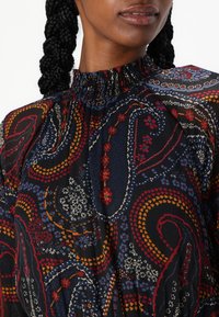 Blusa preta com padrão, apresentando um alto decote franzido, intrincados designs de paisley em vermelho, azul e amarelo, com um tecido texturizado.