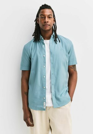 Dreadlockkal rendelkező férfi világoskék rövid ujjú, gombos inget visel, fehér póló fölött, bézs nadrágban, fehér fal mellett áll.