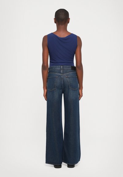 Eine Person, von hinten betrachtet, trägt ein ärmelloses dunkelblaues Oberteil und hochtaillierte, weite, dunkelblaue Jeans vor einem schlichten Hintergrund.