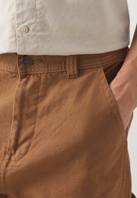 I pantaloni di cotone marroni presentano una chiusura con bottone con logo "Lee", un design a gamba dritta e tasche laterali, mettendo in mostra un tessuto texturizzato.