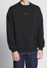 Uomo che indossa una felpa nera con un piccolo logo arancione in stile firma sul petto e jeans blu, con le mani rilassate lungo i fianchi.