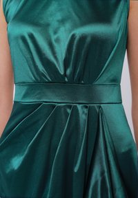 Smaragdgrön satin klänning med en figursydd bodice, plisserad kjol och en texturerad midjeresår. Tyget har en glänsande yta.