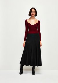 Top a maniche lunghe bordeaux con scollatura profonda a V, abbinato a una gonna maxi nera a coste fluente e stivali neri al ginocchio.