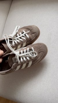 Brune semskede joggesko med hvite striper og snørebånd som hviler på en lysegrå tekstiloverflate, holdt av en hånd.