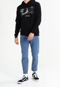 Svart huva sweatshirt med en grafisk framdesign, ihop med ljusblå jeans och svarta sneakers med vita detaljer. Avslappnad passform och stil.