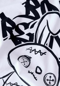 Biała koszulka z czarnym nadrukiem przedstawiającym szytą lalkę z guzikami jako oczami oraz wyboldowanym, stylizowanym tekstem w kolorze czarnym. Miękka tkanina bawełniana.