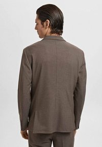 Man met glad achterover gekamd haar, draagt een bruine colbert en bijpassende broek, gezien van achteren tegen een eenvoudige lichte achtergrond.