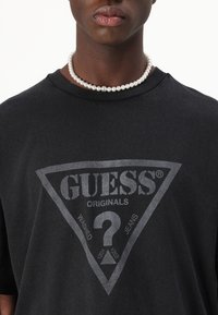Melna kokvilnas T-krekls ar lielu pelēku trīsstūra logo, uz kura ir uzraksts "GUESS ORIGINALS" un jautājuma zīme. Valk�āts ar pērļu kaklarotu.