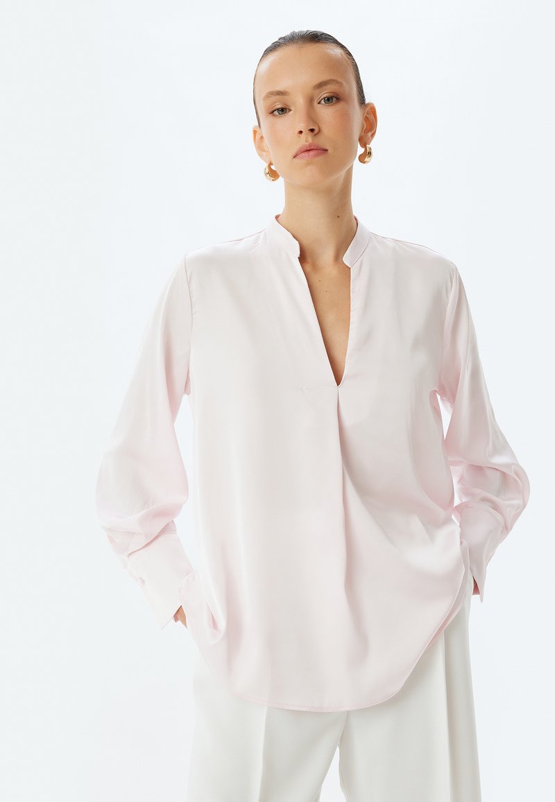 Blusa lunga a maniche e rosa chiaro con colletto a V e dettagli plissettati. Realizzata in tessuto morbido, presenta una vestibilità rilassata e un orlo arrotondato.