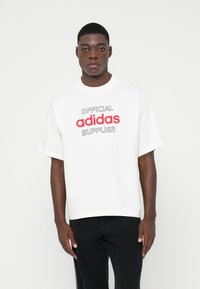 Moški v beli oversize majici s tekstom "Uradni dobavitelj adidas" v rdeči in črni barvi, v kombinaciji s črnimi hlačami, stoji ob bele stene.