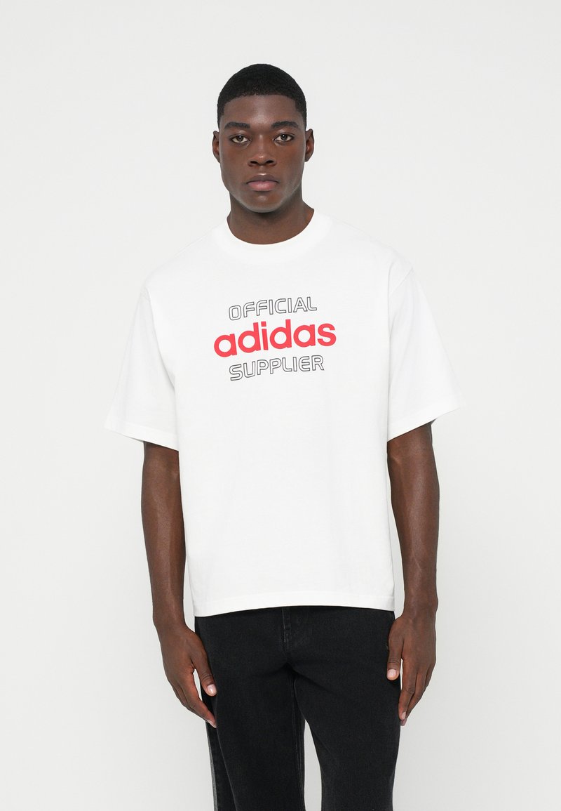 Muž v bielom nadrozmernom tričku s nápisom "Oficiálny dodávateľ adidas" v červenej a čiernej farbe, v kombinácii s čiernymi nohavicami, stojaci na bielom pozadí.