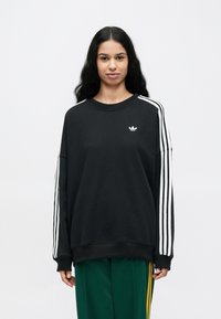 Čierna bavlnená mikina s dlhými rukávmi, na rukávoch sú biele trojité pruhy a na hrudi malá logo.