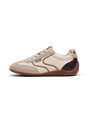Zapatilla beige y marrón con cordones, punta de ante, costuras decorativas y suela marrón texturizada, mostrada de lado sobre fondo blanco.