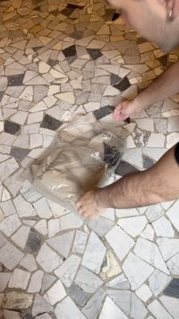 Bežinis drabužis aiškiame plastikinėje maišelyje, kuris atidaromas ant tekstūrinio mozaikos rašto grindų su baltomis, pilkomis ir juodomis plytelėmis.