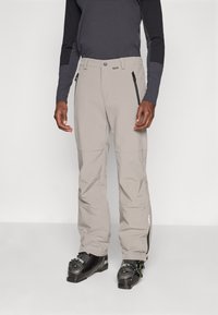 Man wearing gray ski pants with black zipper pockets, black ski boots, and a two-tone long-sleeve top, standing against a plain background.