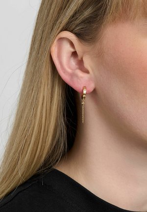 Približen posnetek ušesa osebe s svetlim ravnim blond lasmi, ki nosi majhno zlato obročko z nežno padajočo verižico in drobnim okrasnim obeskom.