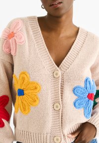 Knitted cardigan in soft beige with oversized, colorful flowers in pink, red, yellow, and blue. Features a deep V-neck and large buttons.