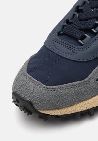 Scarpa da ginnastica blu navy con tomaia in camoscio e tessuto, punta grigia texturizzata, lacci neri piatti e suola in gomma nera robusta.