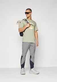 Lys grøn t-shirt med grafisk design, grå tapered bukser med sorte nederste paneler, grå vandrestøvler og en beige rygsæk med sorte accenter.