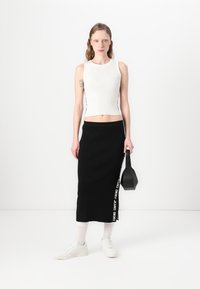 Hvit ribbet crop topp, svart strikket maxiskjørt med sidelogo, hvite sneakers og en liten svart håndveske, stående mot en hvit bakgrunn.