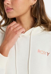 Krémová mikina s kapucňou a predným vreckom, so štruktúrovanou tkaninou, sťahovacími šnúrkami a vyšívaným logom "ROXY" v gradientnej ružovej farbe.