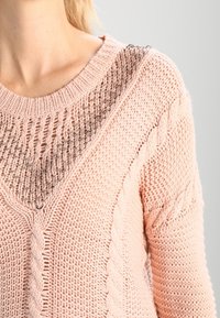 Maglione lavorato a maglia rosa chiaro con un ampio scollo a V testurizzato e dettagli intrecciati sulle maniche. Filato morbido e spesso con un motivo a maglia aperta.