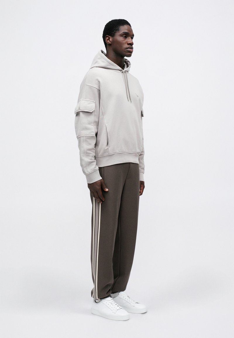 Man standing wearing light beige hoodie with flap pockets on sleeves, dark brown pants with light side stripes, and white sneakers.