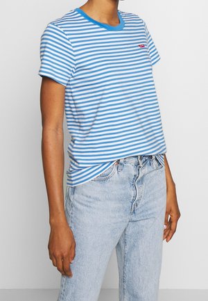 Person iført blå og hvide vandrette stribede kortærmede skjorte og lyseblå højtaljede jeans mod en ensfarvet baggrund.