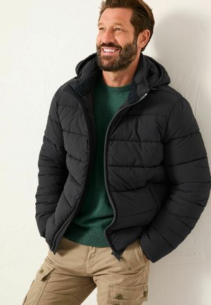 Schwarze Daunenjacke mit Kapuze, Reißverschluss und Fronttaschen. Getragen über einem grünen Pullover, kombiniert mit beigen Cargo-Hosen.