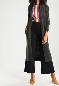 Długi, ciemnozielony kardigan o rypsowanym wzorze, z otwartym przodem i kieszeniami, noszony na paskowanym, wielokolorowym topie oraz czarnych szerokich spodniach.